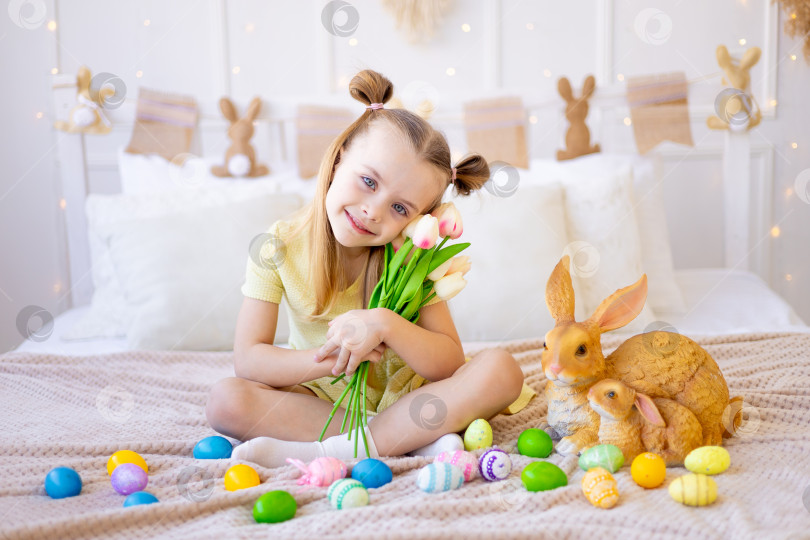 Скачать пасха, маленькая девочка с раскрашенными крашеными яйцами и кроликом, держащими весенние цветы тюльпаны, дома в светлой комнате готовится к празднику, улыбается и веселится фотосток Ozero