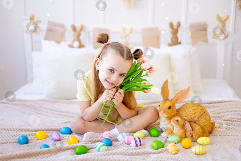 Скачать пасха, маленькая девочка с раскрашенными крашеными яйцами и кроликом, держащими весенние цветы тюльпаны, дома в светлой комнате готовится к празднику, улыбается и веселится фотосток Ozero