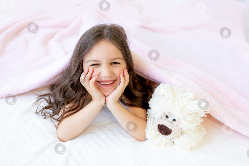 Скачать маленькая улыбающаяся девочка 5-6 лет лежит в постели с плюшевым мишкой, подложив руки под щеки фотосток Ozero
