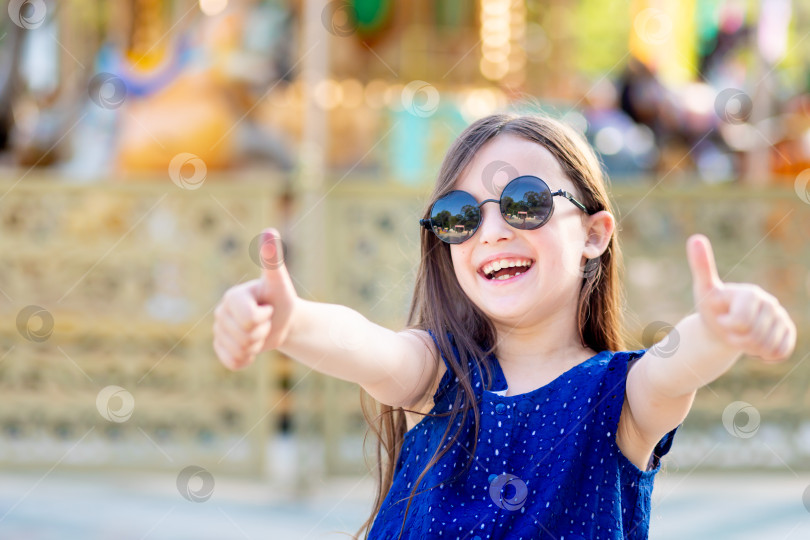 Скачать девочка-ребенок в парке развлечений летом показывает классу поднятый вверх верхний палец и счастливо улыбается каруселям в солнцезащитных очках - концепция летних каникул и школьных каникул фотосток Ozero