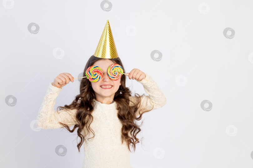 Скачать Симпатичная маленькая девочка с двумя большими леденцами на глазах в праздничной шляпке на белом изолированном фоне с серпантином. Место для текста. Маленькая девочка празднует день рождения, концепция праздника фотосток Ozero