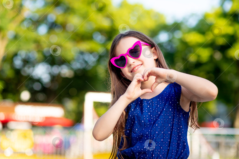 Скачать девочка-ребенок в парке развлечений летом показывает сердечко из своих рук и улыбается от счастья возле каруселей в солнцезащитных очках, концепция летних каникул и школьных каникул фотосток Ozero