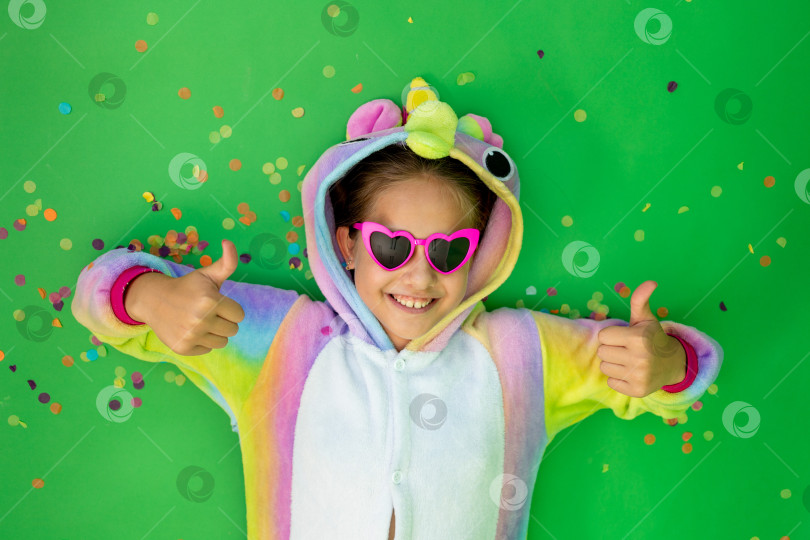 Скачать маленькая девочка в ярком костюме лежит на зеленом изолированном фоне с серпантином и в ярком костюме единорога показывает класс. Место для текста. Концепция праздника, Дня рождения и распродаж фотосток Ozero