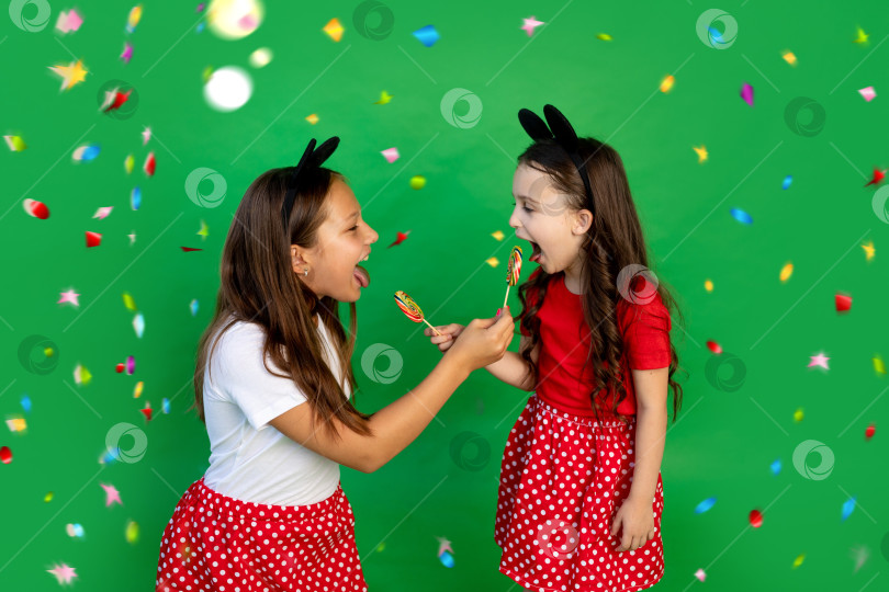 Скачать Милые маленькие девочки в красных нарядах с Микки Маусом на зеленом фоне с серпантином едят большие леденцы на палочке и показывают свои язычки. Пространство для текста.концепция праздника фотосток Ozero