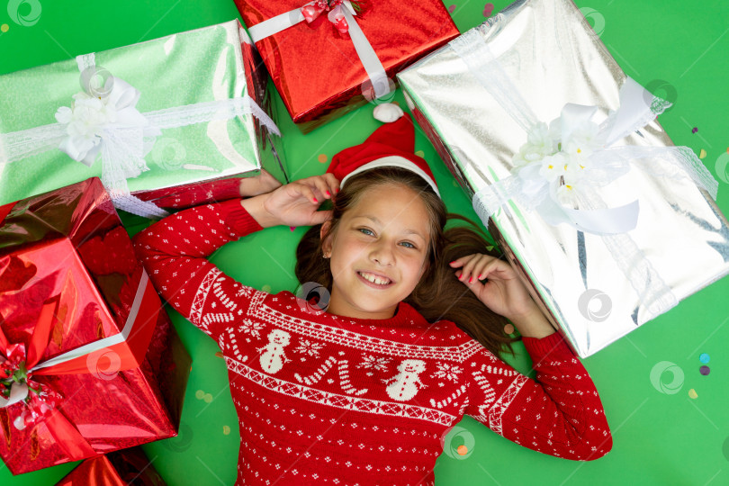Скачать маленькая девочка лежит на зеленом изолированном фоне в красной куртке и шапке Санты среди груды подарков. Место для текста. Концепция нового года и Рождества фотосток Ozero