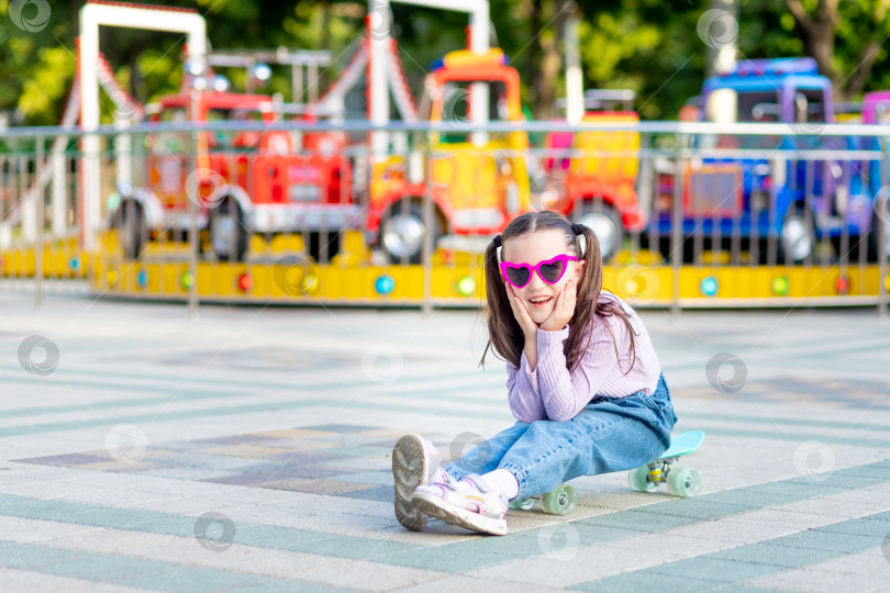 Скачать девочка-ребенок в парке развлечений летом катается на скейтборде и счастливо улыбается возле каруселей в солнцезащитных очках - концепция летних каникул и школьных каникул фотосток Ozero
