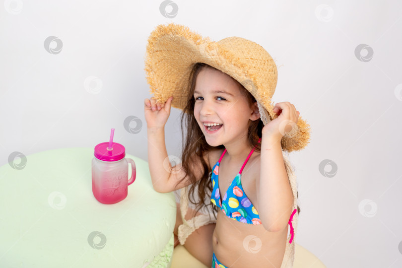 Скачать маленькая улыбающаяся девочка 5-6 лет сидит в купальнике и шляпке с коктейлем на белом изолированном фоне, место для текста фотосток Ozero