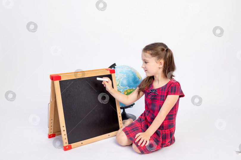 Скачать маленькая школьница 7 лет сидит в красном платье у доски и пишет мелом на белом изолированном фоне, место для текста, 1 сентября, День знаний фотосток Ozero