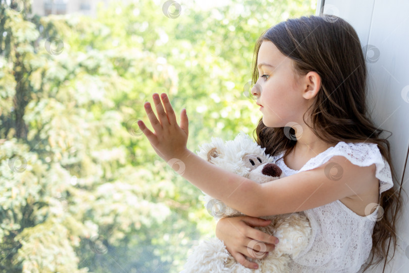 Скачать Очаровательная маленькая девочка 5-6 лет обнимает плюшевого мишку и смотрит в окно фотосток Ozero