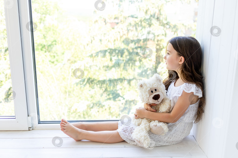 Скачать Очаровательная маленькая девочка 5-6 лет обнимает плюшевого мишку и смотрит в окно фотосток Ozero