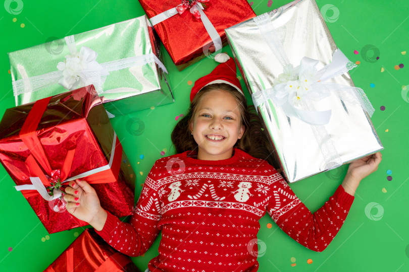 Скачать маленькая девочка лежит на зеленом изолированном фоне в красной куртке и шапке Санты среди груды подарков. Место для текста. Концепция нового года и Рождества фотосток Ozero