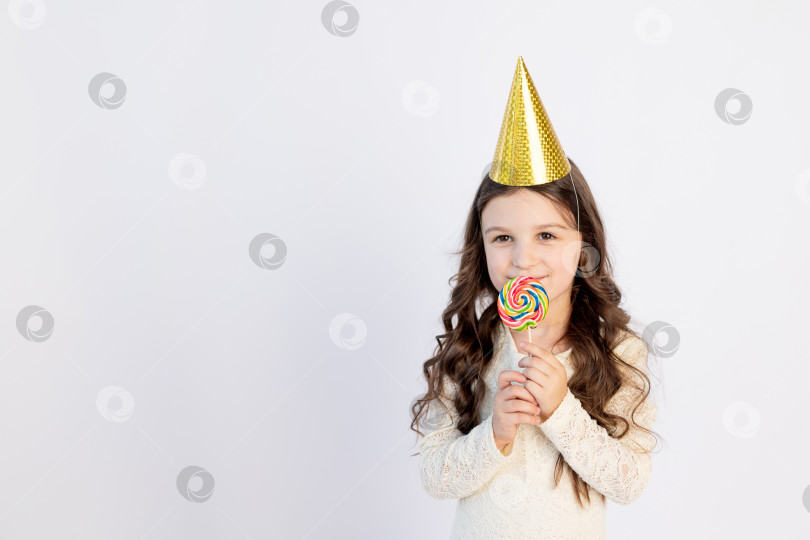 Скачать Милая маленькая девочка ест большой леденец на палочке в праздничной шляпке на белом изолированном фоне. Место для текста. Маленькая девочка празднует день рождения, концепция праздника фотосток Ozero