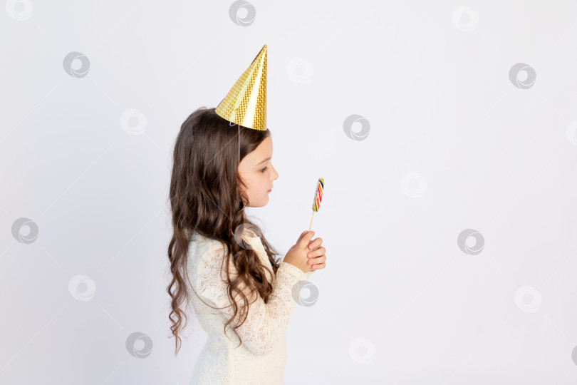 Скачать Милая маленькая девочка ест большой леденец на палочке в праздничной шляпке на белом изолированном фоне. Место для текста. Маленькая девочка празднует день рождения, концепция праздника фотосток Ozero