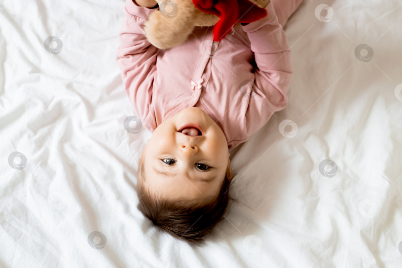 Скачать портрет улыбающейся маленькой девочки, лежащей на кровати с плюшевым мишкой. пробел для текста фотосток Ozero