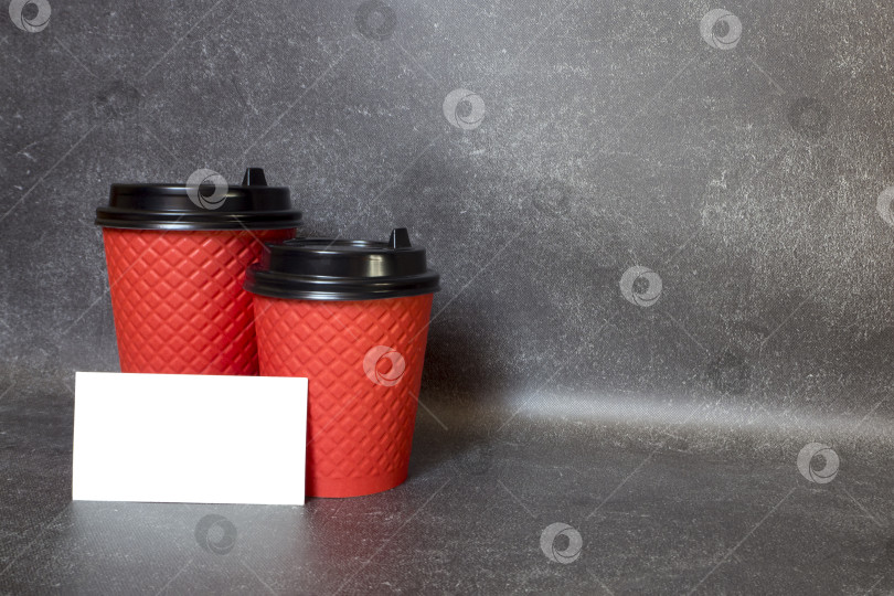 Скачать Две красные одноразовые кофейные чашки с черными пластиковыми крышками и пустая белая визитная карточка на темно-сером бетонном фоне. Концепция "Кофе в дорогу". Горизонтальное изображение при естественном освещении, пространство для копирования, макет. фотосток Ozero