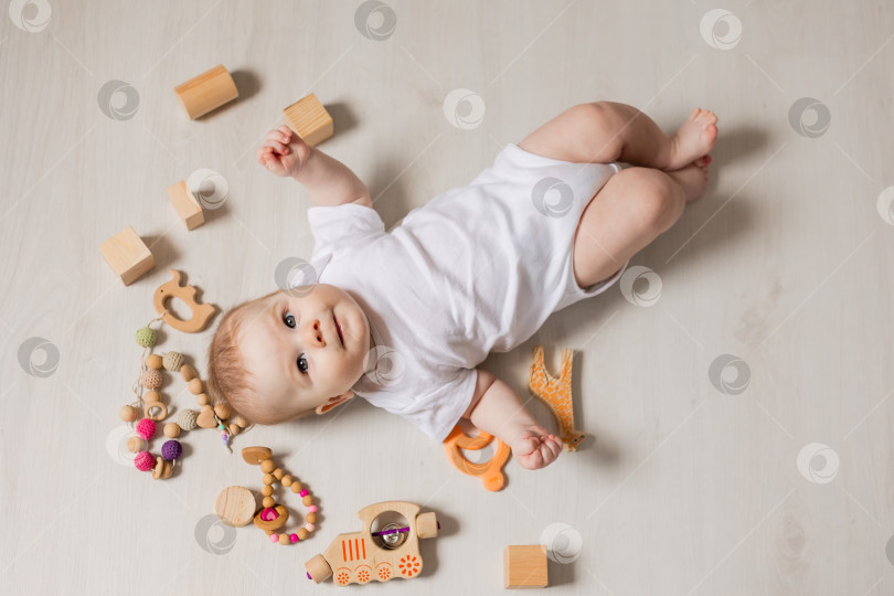 Скачать милый малыш в белом боди лежит на деревянном полу среди деревянных развивающих игрушек фотосток Ozero