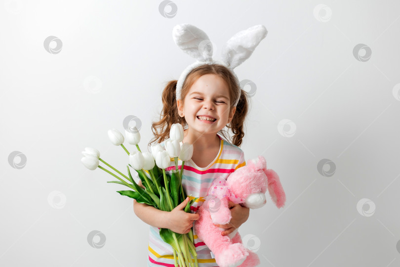 Скачать портрет милой девушки на светлом фоне, с заячьими ушками на голове, с мягкой игрушкой в руках и тюльпанами, концепция Пасхи фотосток Ozero
