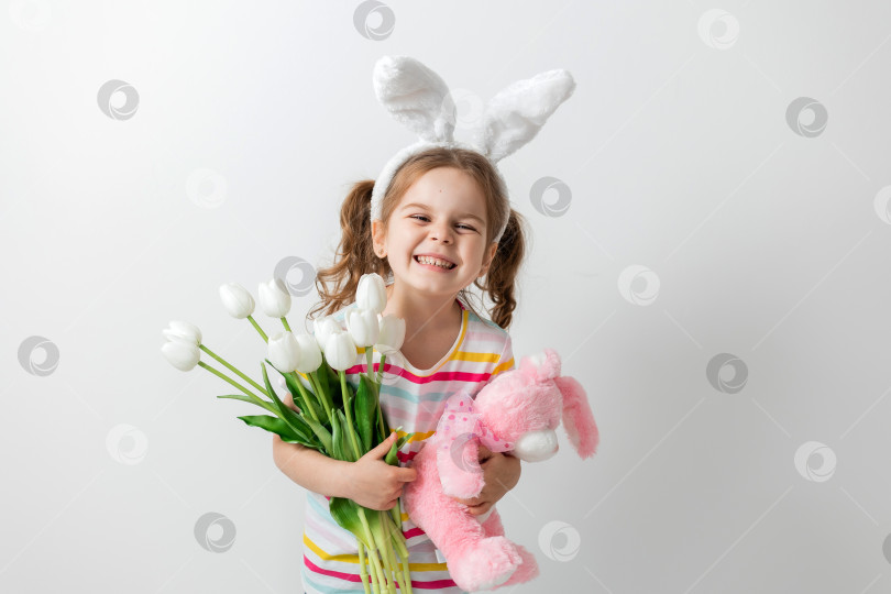 Скачать портрет милой девушки на светлом фоне, с заячьими ушками на голове, с мягкой игрушкой в руках и тюльпанами, концепция Пасхи фотосток Ozero