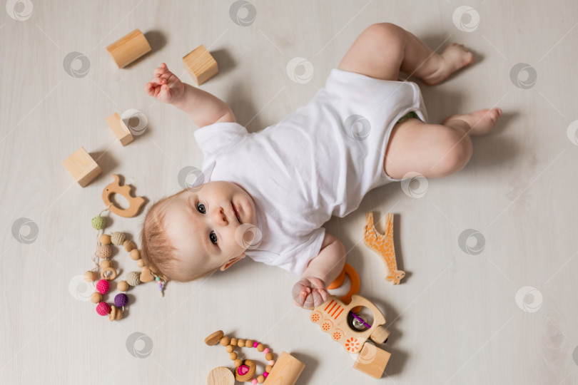 Скачать милый малыш в белом боди лежит на деревянном полу среди деревянных развивающих игрушек фотосток Ozero