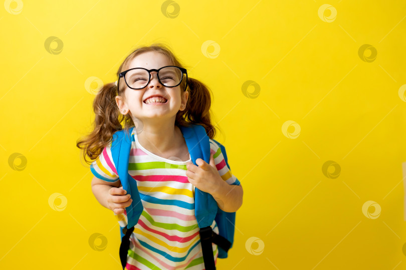 Скачать маленькая девочка в цветной футболке и очках, держащая школьные тетради и рюкзак за плечами на желтом фоне фотосток Ozero