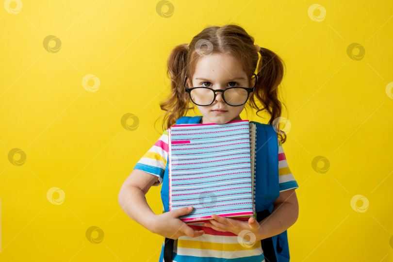 Скачать маленькая девочка в цветной футболке и очках, держащая школьные тетради и рюкзак за плечами на желтом фоне фотосток Ozero