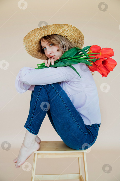 Скачать Молодая женщина держит в руках букет красных тюльпанов и сидит на деревянном стуле. Концепция 8 марта, Дня святого Валентина. Замечательный подарок для девушки. Весенний портрет женщины в соломенной шляпе. фотосток Ozero