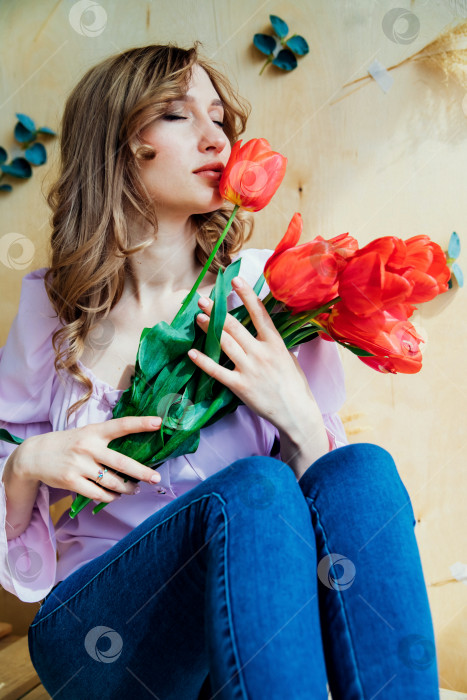 Скачать Молодая женщина держит в руках букет красных тюльпанов и сидит на деревянном подиуме. Концепция 8 марта, Дня святого Валентина. Замечательный подарок для девушки. Весенний портрет женщины. фотосток Ozero