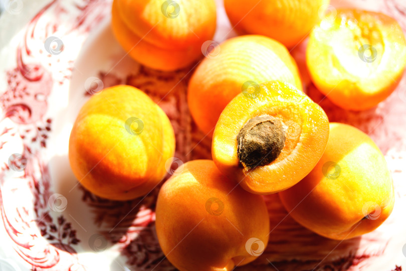 Скачать Спелые сырые абрикосы в тарелке на столе в солнечный день, крупным планом, вертикально. Горка свежих цельных сочных абрикосов оранжевого цвета и половинок абрикоса с косточкой на тарелке из английского фарфора, вид сверху. фотосток Ozero