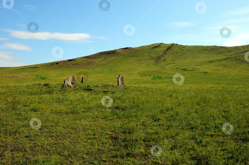 Скачать Древние каменные менгиры на широкой поляне, заросшей высокой травой, у подножия высокой горы с параллельными каменными образованиями. фотосток Ozero