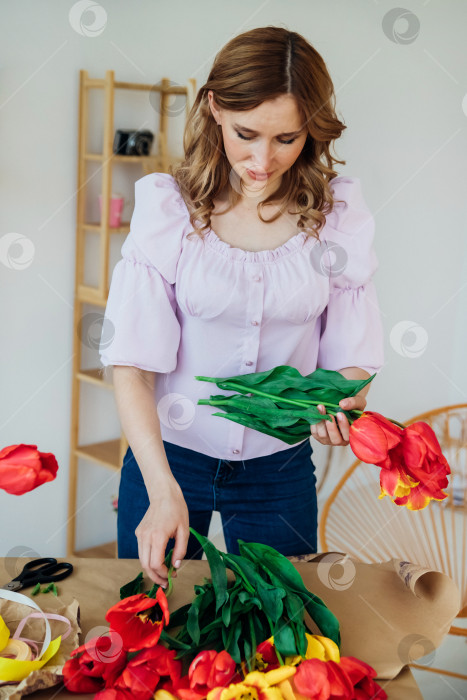 Скачать Молодая женщина упаковывает праздничный букет в оберточную бумагу. Флорист делает сборку из красных тюльпанов в мастерской. Женщина на работе, в малом бизнесе или хобби. фотосток Ozero