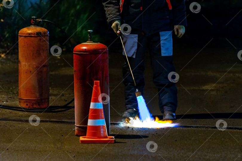 Скачать Пламя газовой горелки в выборочном фокусе во время ямочного ремонта дороги нагревает асфальтовое покрытие, подготавливая дорогу к нанесению слоя битума. фотосток Ozero