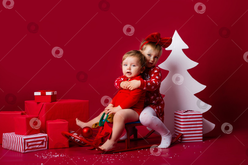 Скачать двое детей сидят на санках с рождественскими подарками в студии на красном фоне. Рождественская концепция, текстовое пространство фотосток Ozero