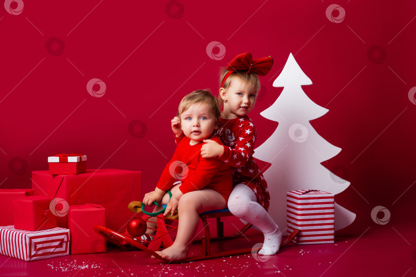 Скачать двое детей сидят на санках с рождественскими подарками в студии на красном фоне. Рождественская концепция, текстовое пространство фотосток Ozero