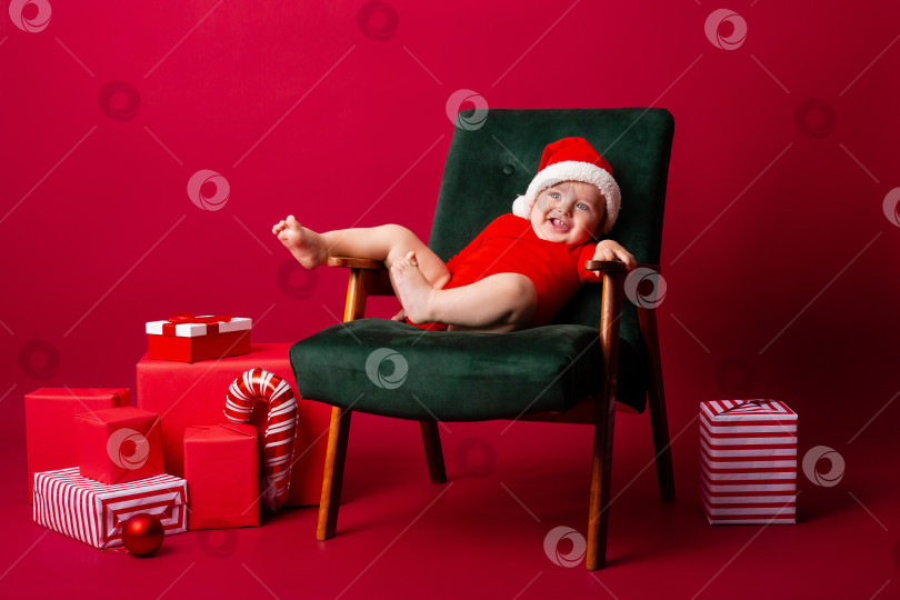 Скачать Милый малыш Санта сидит на стуле среди рождественских подарков на красном фоне. Рождественская концепция, текстовое пространство фотосток Ozero