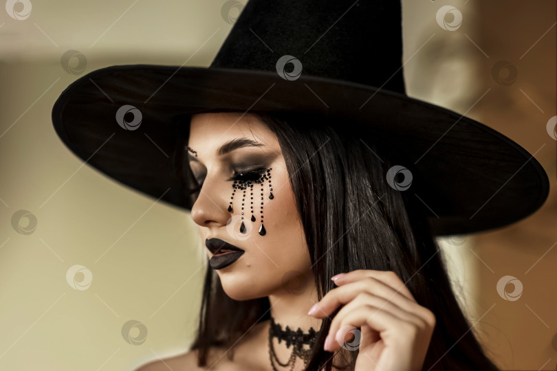 Скачать Молодая девушка в образе ведьмы, в черной шляпе и черном платье с макияжем в готическом стиле выглядит как ведьма, концепция идей Хэллоуина фотосток Ozero