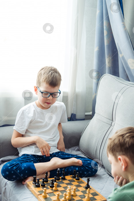 Скачать Мальчики играют в деревянные шахматы, лежа на сером диване перед окном фотосток Ozero