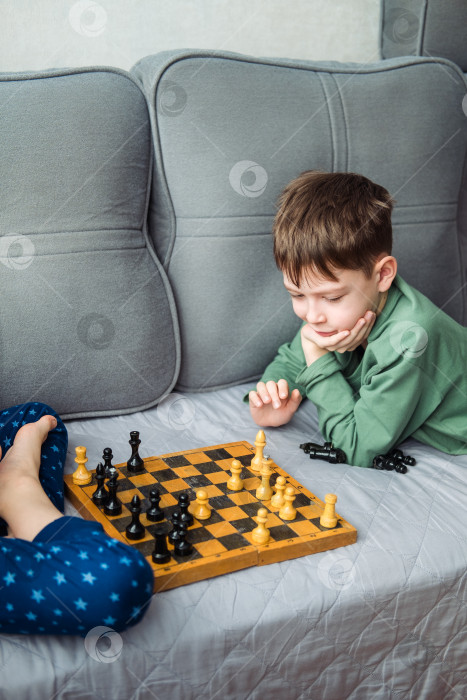 Скачать Мальчики играют в деревянные шахматы, лежа на сером диване. фотосток Ozero