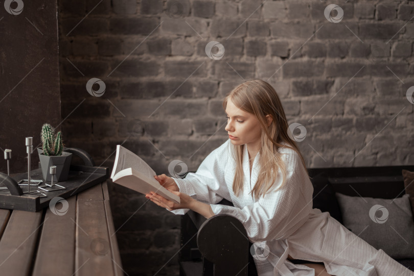 Скачать Молодая красивая блондинка в белом халате сидит у окна на диване и читает бумажную книгу. фотосток Ozero