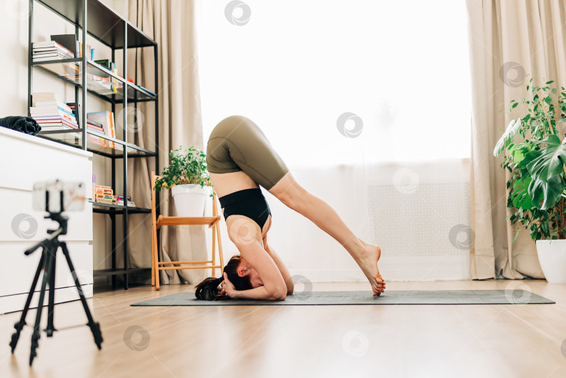 Подтянутая женщина тренируется, выполняя упражнения на растяжку на коврике для йоги во время видеотрансляции фитнес-видео онлайн на смартфоне на штативе дома. Женщина, практикующая упражнение йоги онлайн - Ozero - российский фотосток