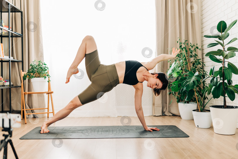 Скачать Подтянутая женщина тренируется, выполняя упражнения на растяжку на коврике для йоги во время видеотрансляции фитнес-видео онлайн на смартфоне на штативе дома. Женщина, практикующая упражнение йоги онлайн фотосток Ozero