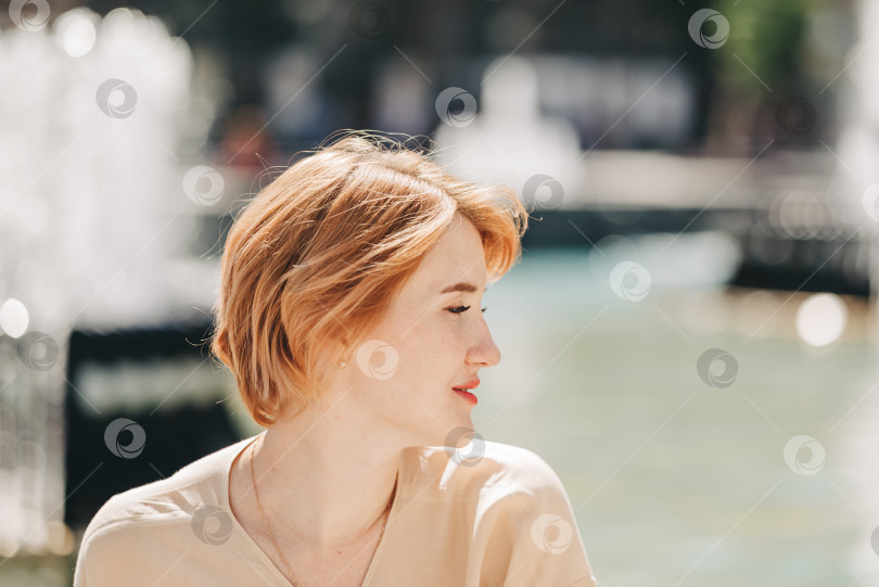 Скачать Молодая красивая женщина с рыжими короткими волосами сидит на открытом воздухе в городском парке возле фонтана. Летний солнечный день фотосток Ozero