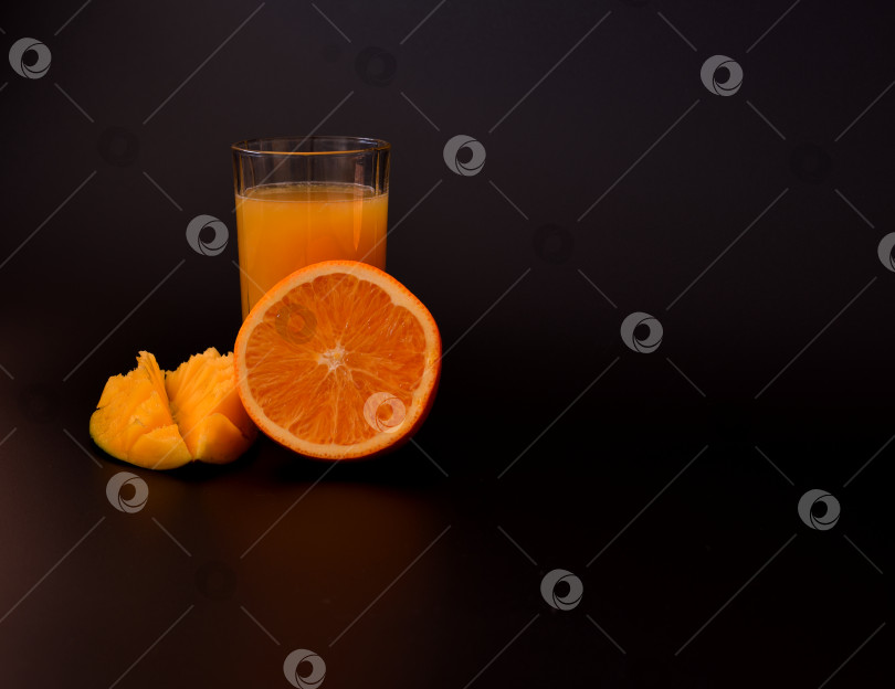 Скачать Стакан смеси свежевыжатых фруктовых соков на черном фоне, рядом с кусочками спелого манго и половинкой апельсина. фотосток Ozero