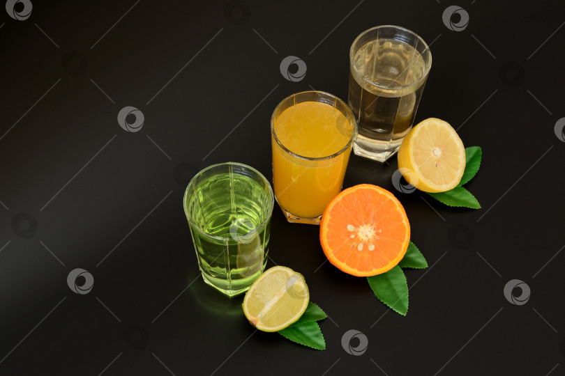Скачать Три высоких бокала с разными цитрусовыми соками на черном фоне, рядом с половинками спелого апельсина, лимона и лайма с листьями. фотосток Ozero