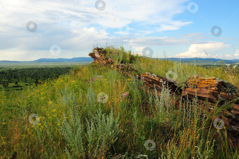 Скачать Слоистое скальное образование из красного песчаника на вершине высокого травянистого холма с видом на живописную долину в солнечный летний день. фотосток Ozero