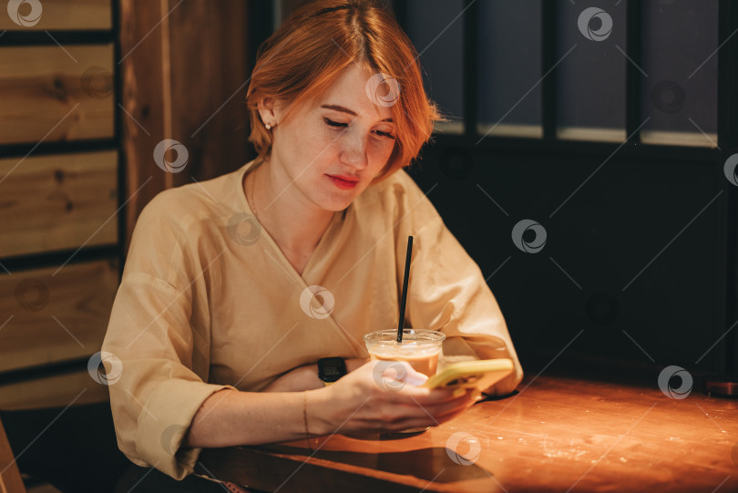 Скачать Молодая женщина использует смартфон для видеочата и общения онлайн с Wi-Fi Интернетом в кафе с низким освещением cozy vibes cafeteria. Стиль жизни с короткой стрижкой женского рыжего цвета. Звонок по мобильному телефону фотосток Ozero