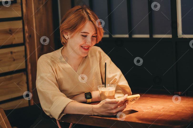 Скачать Молодая женщина использует смартфон для видеочата и общения онлайн с Wi-Fi Интернетом в кафе с низким освещением cozy vibes cafeteria. Стиль жизни с красной короткой стрижкой. Звонок по мобильному телефону фотосток Ozero