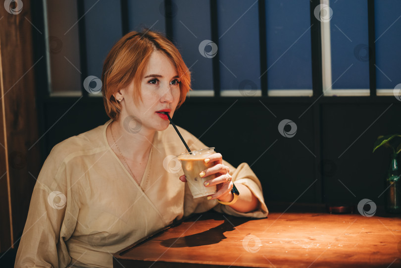 Скачать Женщина, пьющая кофе со льдом капучино или латте в кафе, сидя рядом за столиком при слабом освещении, темная фотография образа жизни. Красивая уверенная в себе женщина с короткими рыжими волосами одна в кафетерии выпивает фотосток Ozero