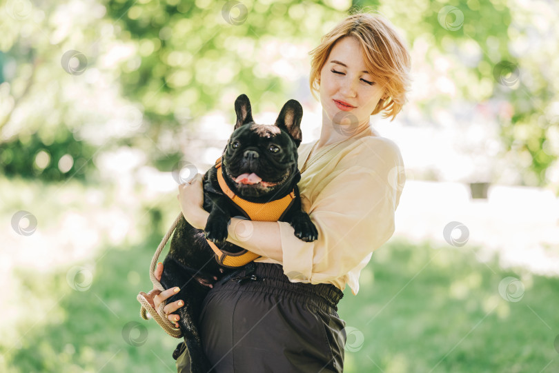 Скачать Щенок черного французского бульдога обнимается с девушкой-хозяйкой, лежащей на лужайке в парке на открытом воздухе летним весенним днем, целующейся, обнимающейся, ласкающейся. Счастливая женщина с собакой на улице фотосток Ozero