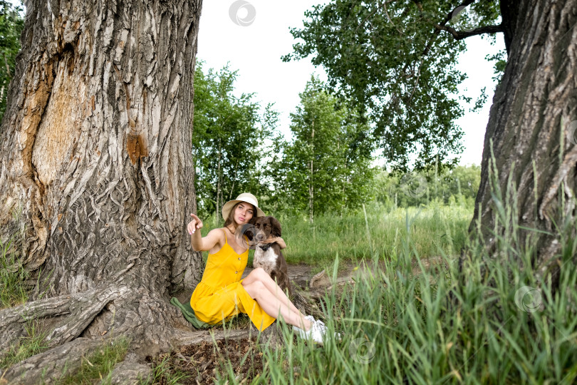 Скачать Симпатичная молодая женщина в желтом платье и шляпе сидит под деревом с собакой. Женщина расслабляется в жаркий летний день. фотосток Ozero