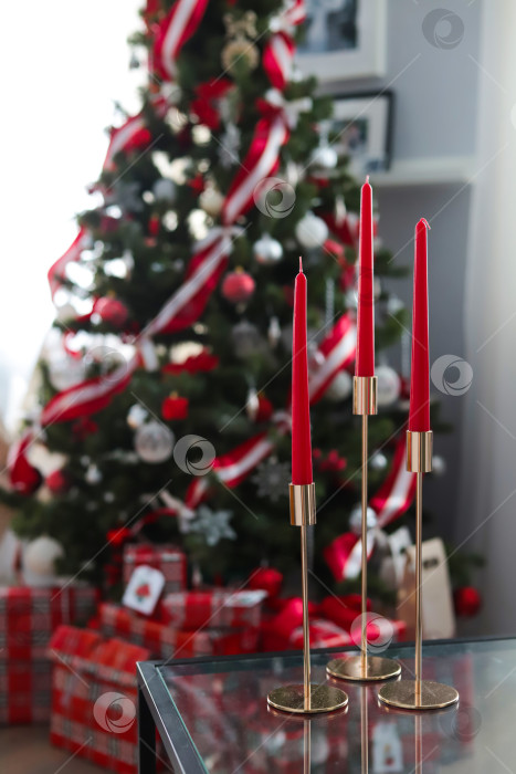 Скачать Красные свечи в золотых подсвечниках на фоне рождественской елки, украшенной красными игрушками и атласными лентами. Подарки под елкой. фотосток Ozero